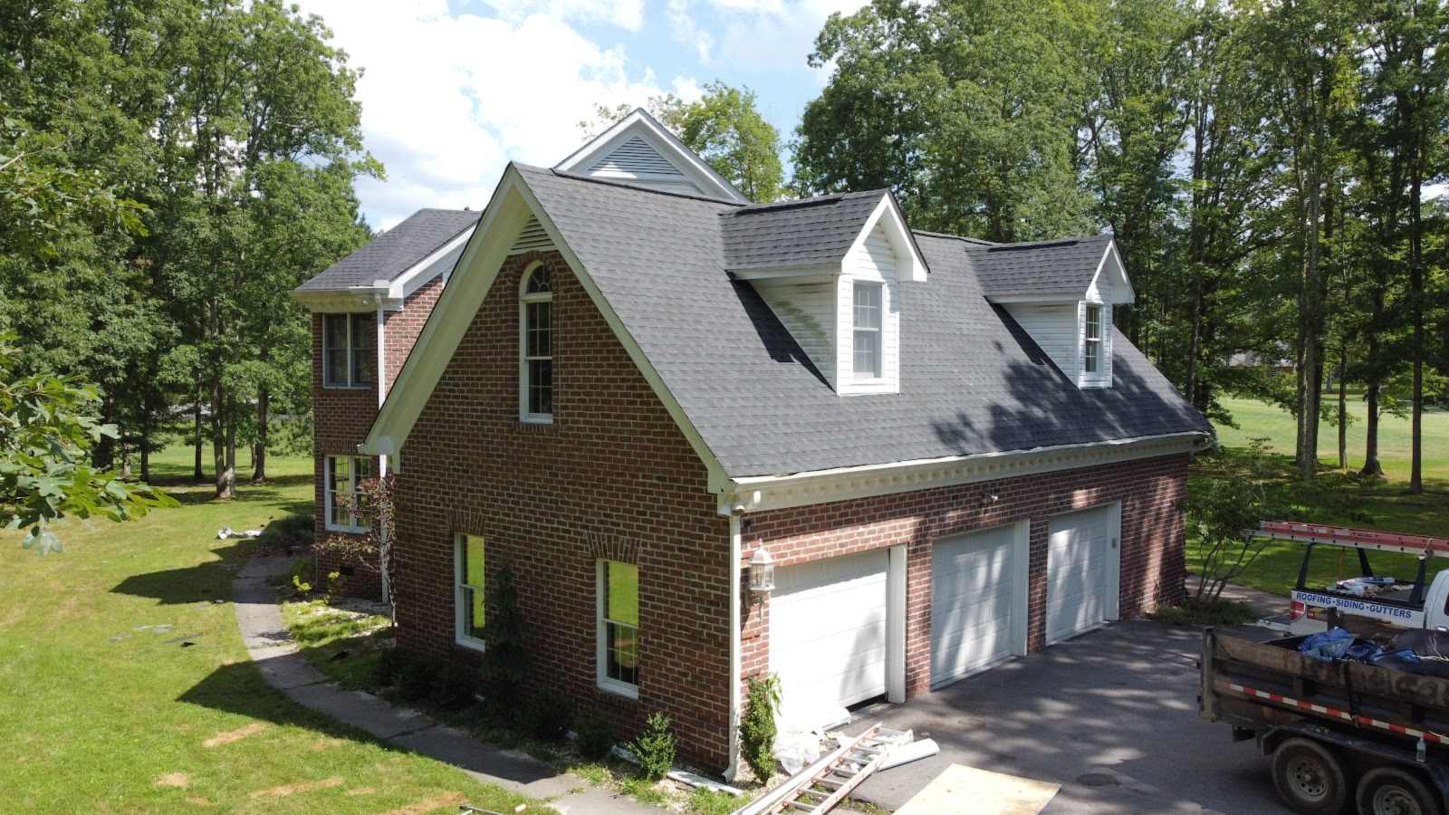 Drone view of a red-brick house with a new gray shingle roof, two dormers and a three-car garage, trees on the left and a work trailer in the drive.