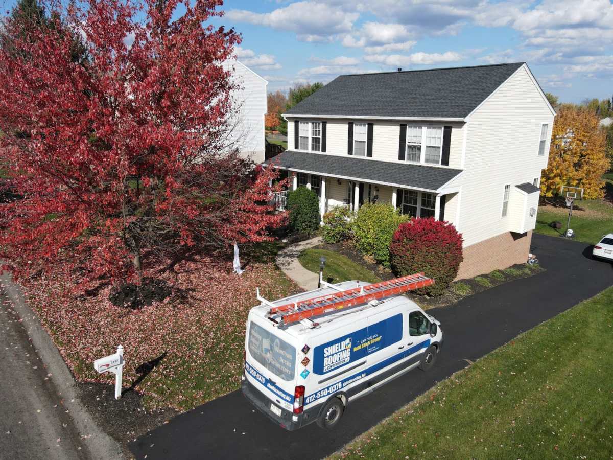 Drone view of a white Shield Roofing van with a ladder on the roof rack, parked in a blacktop driveway beside a lawn and red shrubs.