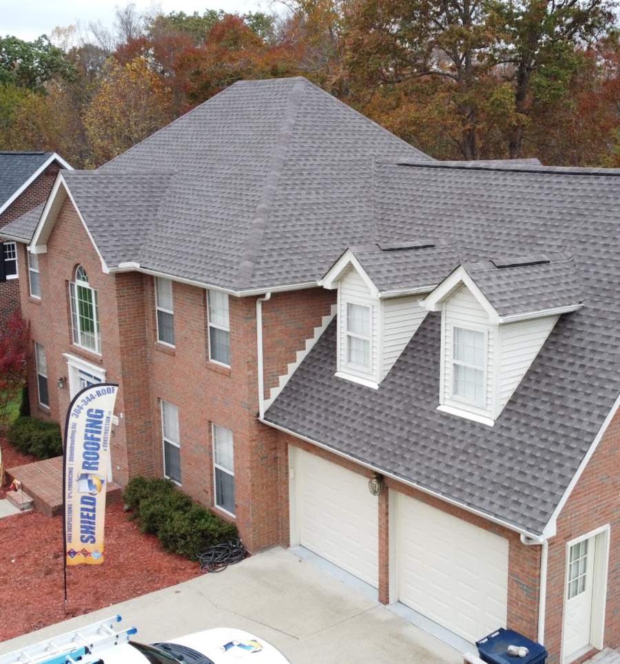 Drone view of a brick two-story house with a new gray roof and dormers, a blue and gold Shield Roofing feather flag planted by the front walk.
