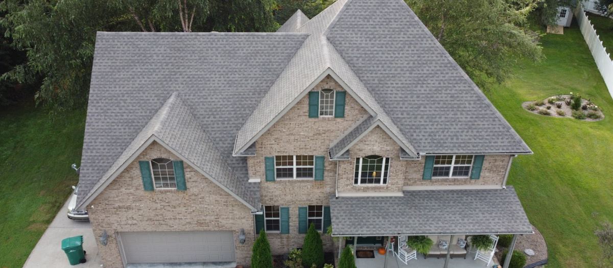 Drone view from above of a large tan-brick house with a new gray shingle roof, green shutters and a landscaped front yard.